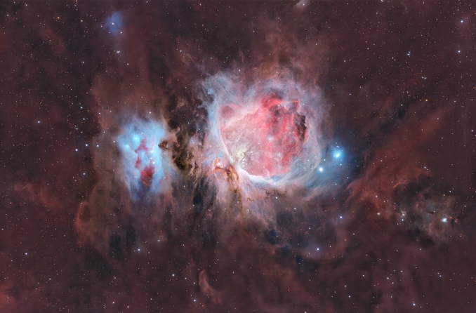 Nel grande respiro della regione di Orione, M42 non è sola: è un cuore luminoso immerso in un paesaggio più vasto, dove vapori cremisi e veli azzurri si dissolvono lentamente nellombra. - Copyright: Massimo Botti & Giuseppe Capriotti
